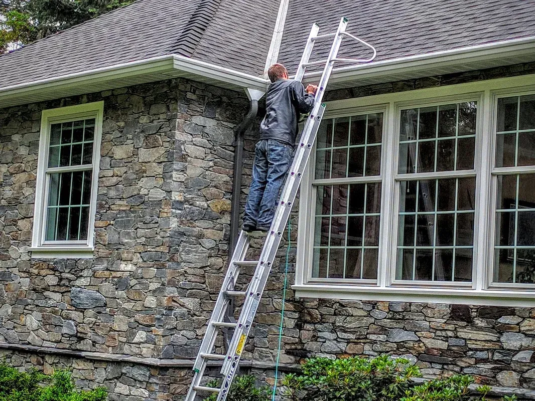 A worker up an extension ladder at the roof edge of a stone house